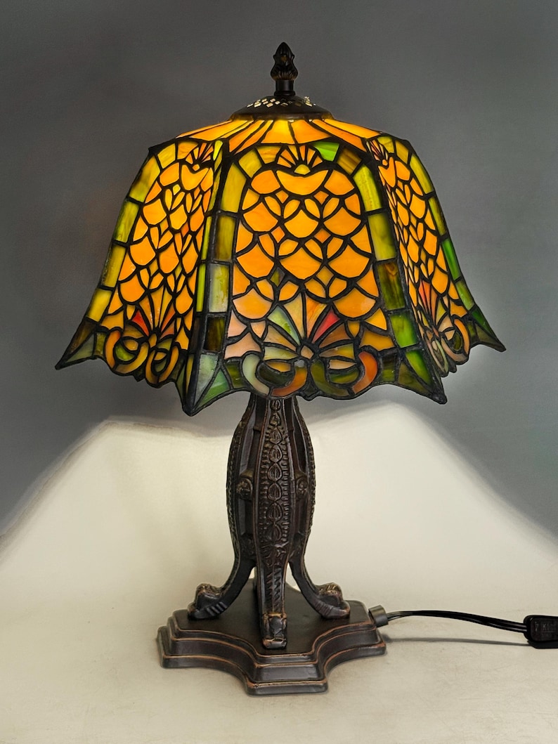 Op de afbeelding: Een glas-in-lood tafellamp met een uitlopende kap voorzien van een geometrisch schubpatroon in levendige oranje en groene tinten. De lamp heeft een donkere, getextureerde metalen voet met een decoratieve steel en een vierkante voet.