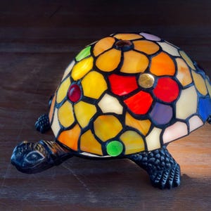 Może przedstawiać: Lampa w kształcie żółwia ze witrażu z kolorową muszlą mozaikową. Muszla ma żywą mieszankę żółtych, pomarańczowych, czerwonych, fioletowych i niebieskich szklanych elementów. Ciało i nogi żółwia są ciemne.