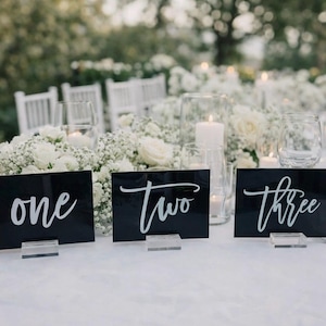May include: A beautifully decorated table setting with white flowers, candles, and place settings. Black table number cards with white script read "one", "two", and "three". The scene is set outdoors with soft, natural lighting.