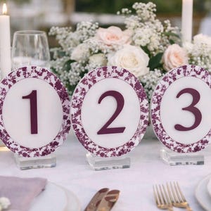 May include: Elegant table setting with three decorative table number cards. Each card features a white background with a burgundy floral border and a large burgundy number. The table is decorated with flowers and candles.