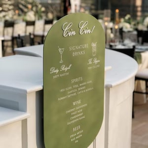 May include: A green, arched sign with white text and illustrations, listing signature drinks, spirits, wine, and beer. The sign reads "Cin Cin!" and includes drink names like "Dirty Martini" and "Tom Collins."