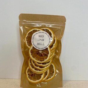 May include: A clear plastic bag filled with dried lemon wheels. The bag is sealed and has a brown paper backing. A white circular label with the text "Dried Lemon Wheels" is visible on the front of the bag. The dried lemon slices are a light brown color.