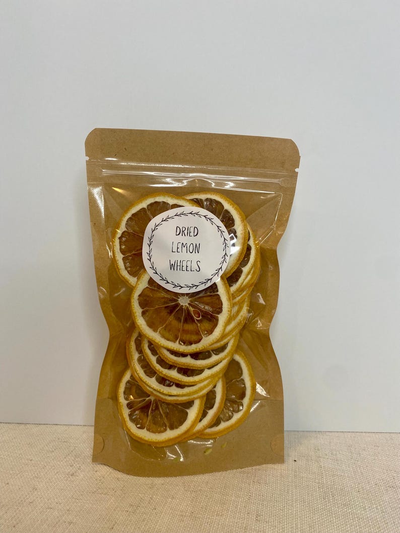 May include: A clear plastic bag filled with dried lemon wheels. The bag is sealed and has a brown paper backing. A white circular label with the text "Dried Lemon Wheels" is visible on the front of the bag. The dried lemon slices are a light brown color.