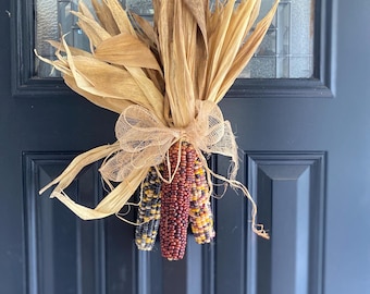 May include: A decorative bundle of three dried multi-colored flint corn cobs with tan husks tied together with a light brown mesh ribbon. The corn kernels display shades of deep red, yellow, orange, and dark blue, arranged in a vertical cluster.
