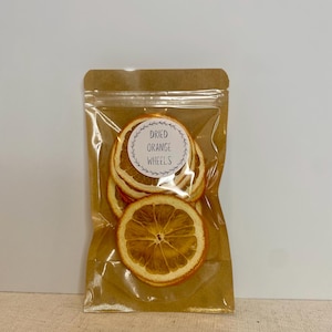 May include: A clear plastic bag filled with dried orange wheels. The bag is sealed and attached to a brown paper backing. The label on the bag reads "Dried Orange Wheels" in a circular design.