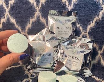 May include: Four silver foil packets containing round, light green shower steamers with the text "REFRESH shower steamer EUCALYPTUS, MINT ROSEMARY & MENTHOL" printed on the front of each packet.