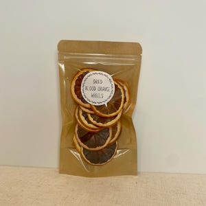 May include: A clear plastic bag filled with dried blood orange wheels. The bag is sealed and has a brown paper backing. A white circular sticker with the words "Dried Blood Orange Wheels" is affixed to the front of the bag.