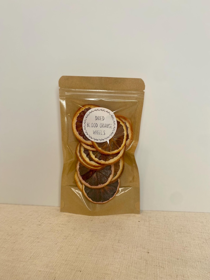 May include: A clear plastic bag filled with dried blood orange wheels. The bag is sealed and has a brown paper backing. A white circular sticker with the words "Dried Blood Orange Wheels" is affixed to the front of the bag.