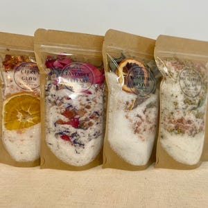 May include: Four clear pouches filled with bath salts. Each pouch has a different blend, including Citrus Glow, Lavender Lullaby, Rosemary Revival, and Eucalyptus Escape. The pouches are sealed and contain dried flowers and citrus slices.