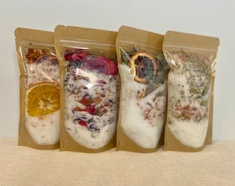 May include: Four clear pouches filled with bath salts. Each pouch has a different blend, including Citrus Glow, Lavender Lullaby, Rosemary Revival, and Eucalyptus Escape. The pouches are sealed and contain dried flowers and citrus slices.