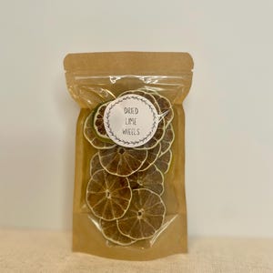 May include: A clear plastic bag filled with dried lime wheels. The bag has a brown paper top and a white label with the words "Dried Lime Wheels" in a circular design. The lime slices are a light brown color.