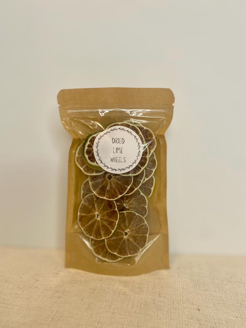 May include: A clear plastic bag filled with dried lime wheels. The bag has a brown paper top and a white label with the words "Dried Lime Wheels" in a circular design. The lime slices are a light brown color.