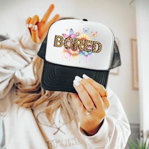 Op de afbeelding: Een witte trucker-pet met een zwart mesh-achterpaneel. De voorkant van de pet heeft een kleurrijke afbeelding van bloemen en vlinders met het woord "BORED" in luipaardprint.