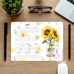 Op de afbeelding: Een rechthoekige bureaumat met een kalender voor 2026 en een zonnebloemontwerp. De kalender is wit met gele en groene bloemaccenten. Een aquarel illustratie van zonnebloemen in een glazen pot staat rechts.