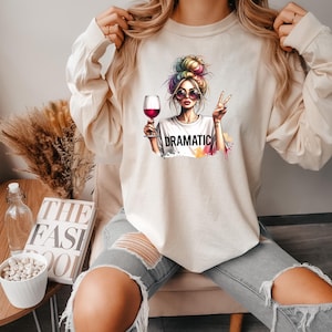 May include: A cream colored long-sleeved sweatshirt with a graphic print of a woman with colorful hair holding a glass of red wine. The text "DRAMATIC" is printed below the woman.