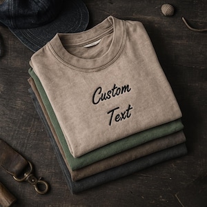 Könnte beinhalten: Ein Stapel von fünf T-Shirts in gedämpften Farben, darunter Beige, Grün und Grau, wobei das oberste Shirt mit den Worten "Custom Text" in Schwarz bestickt ist. Eine dunkelblaue Kappe und ein Lederriemen sind sichtbar.