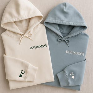 May include: Two hooded sweatshirts, one cream and one blue, are shown. Both have the Roman numeral date "IX.VIIMMXVI" embroidered on the chest. The cream sweatshirt has a crescent moon and heart on the cuff, while the blue one has a "Y" and heart.