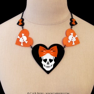 May include: A statement necklace featuring a black heart pendant with a white skull and orange bow. The necklace includes orange heart-shaped charms with white skull and crossbones, and black heart-shaped accents.