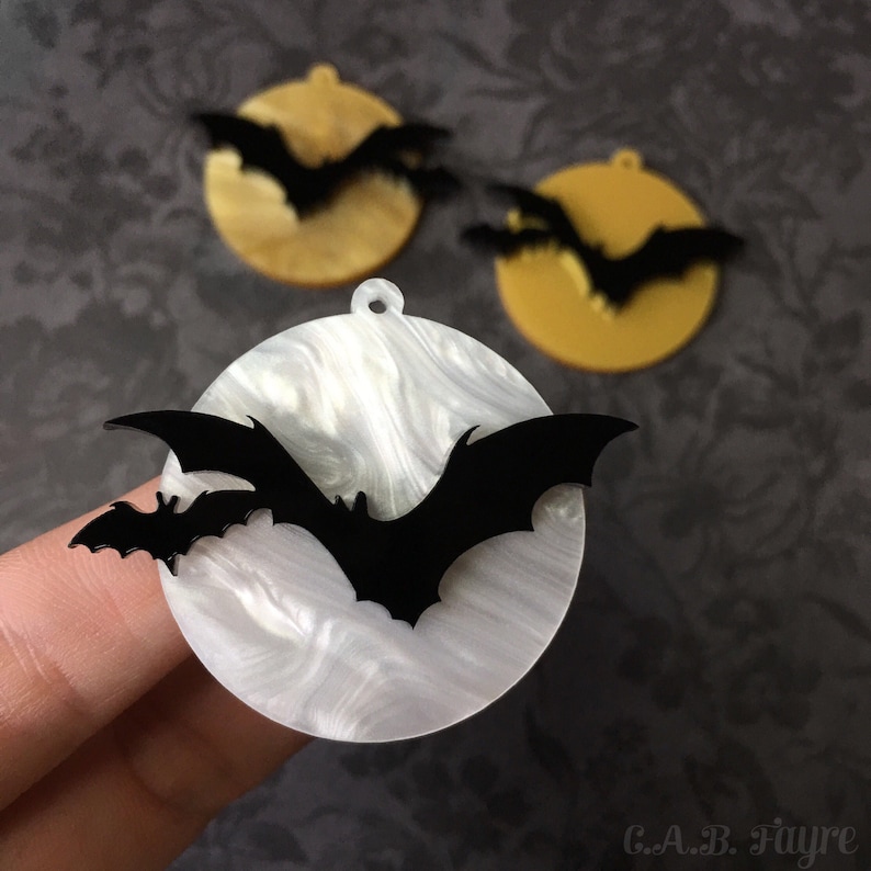 Close-up of white pearl moon bat earring with black bats, ghostly Halloween jewelry with gold moon versions in background.