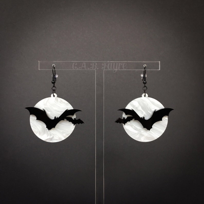 White pearl moon Halloween earrings with layered black bat silhouettes, displayed on clear stand for gothic spooky jewelry.