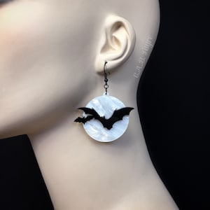 Spooky bat earrings with white pearl moon background and black flying bats, shown on mannequin ear for Halloween costume style.