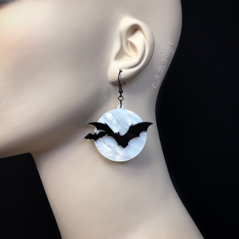 Spooky bat earrings with white pearl moon background and black flying bats, shown on mannequin ear for Halloween costume style.