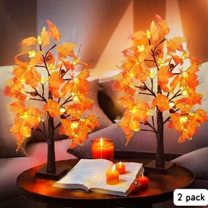 May include: Two decorative LED maple leaf trees with warm white lights. The trees have brown trunks and bases, and orange leaves. The scene includes lit candles, an open book, and small pumpkins on a round table. The text "2 pack" is visible.