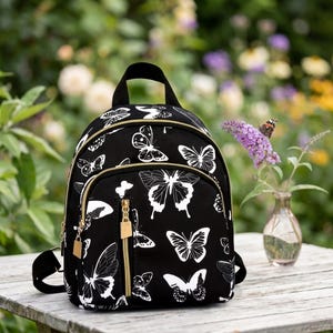 May include: A black backpack featuring a white butterfly pattern, gold-tone zippers, a top handle, and adjustable shoulder straps. The bag includes a main zippered compartment and a smaller front zippered pocket with a rectangular gold zipper pull.