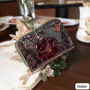 May include: A black leather wallet with a floral design. The wallet features embossed burgundy flowers and a gold zipper. The word "Wallet" is in the bottom right corner.