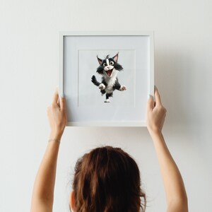 May include: A person hangs a white framed picture on a white wall. The picture features a cartoon black and white cat with a surprised expression.