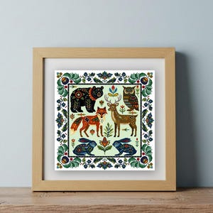 Op de afbeelding: Een ingelijste print met illustraties in volkskunststijl van bosdieren: een beer, een vos, een hert, een uil en konijnen. De dieren zijn omgeven door een bloemenrand. Het kunstwerk wordt gepresenteerd in een lichtbruine houten lijst.