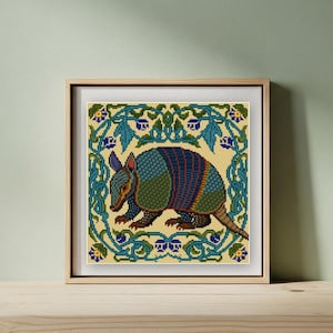 May include: Framed cross-stitch art featuring a detailed armadillo. The artwork showcases the animal in shades of blue, green, and brown, set against a cream background. A border of blue floral patterns surrounds the armadillo, all within a wooden frame.