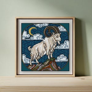May include: Framed cross-stitch art depicting a white goat with large horns on a mountain. The background is dark teal with white clouds and a crescent moon. The artwork is in a wooden frame.