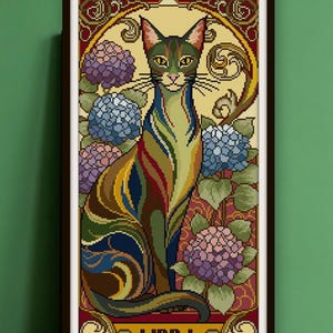 May include: Framed cross-stitch art featuring a stylized cat with colorful striped fur, set against a backdrop of blue and purple hydrangeas. The artwork is framed in a dark brown frame, with the word "LIBRA" at the bottom.