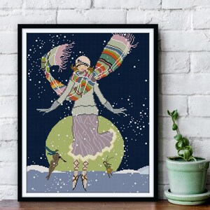 May include: Framed cross-stitch art featuring a woman in a winter scene. The woman wears a hat, scarf, and dress in pastel colors, with a green and blue background. The artwork is in a black frame and is displayed against a white brick wall.