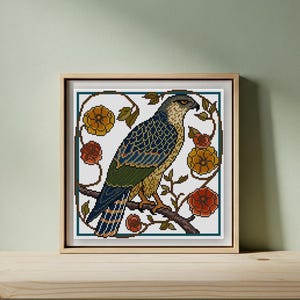 May include: Cross-stitch art of a bird of prey perched on a branch, surrounded by stylized flowers. The bird features blue, green, and gold details. The artwork is framed in a wooden frame and set against a light green background.