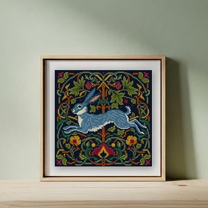 May include: A framed cross-stitch art piece featuring a blue hare leaping across a dark blue background. The background is filled with a floral pattern in green, yellow, red, and orange. The artwork is displayed in a light brown wooden frame.