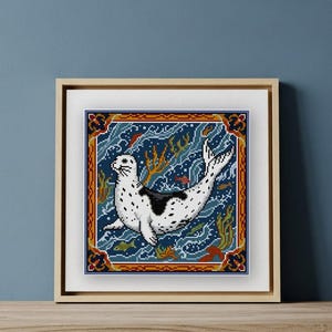 May include: Framed cross-stitch art featuring a white and black spotted seal swimming in blue water with fish and seaweed. The artwork is framed in a light wood frame with a white mat.