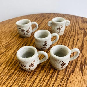 May include: A set of five small, speckled, off-white ceramic mugs with brown hand-painted floral designs. Each mug features a rounded body, a single handle, and a smooth glazed finish. The collection includes one mug with two handles.