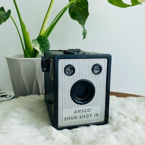 May include: A black Ansco Shur-Shot Jr. box camera featuring a silver textured front panel with two small circular viewfinders and a central lens. The camera body is rectangular with a black carrying handle on the top surface.