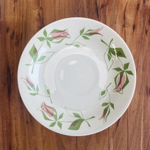 May include: A round white ceramic bowl featuring a repeating pattern of stylized pink rosebuds with green leaves and stems arranged in a circular design around the inner rim. The bowl has a smooth, glossy finish and a simple, clean edge.