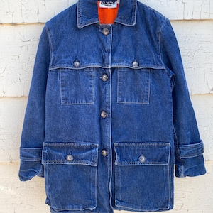 May include: A blue denim jacket with four front pockets and button closures. The jacket has a collared neckline and a bright orange lining. The brand name "DKNY" is visible on the collar. The jacket is hanging on a wooden hanger.