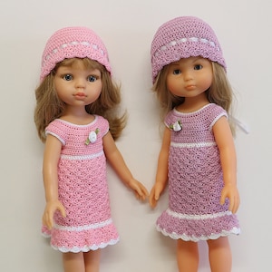 Könnte beinhalten: Zwei Puppen in passenden, handgefertigten Häkel-Outfits. Jede Puppe trägt ein rosa Kleid mit weißer Bordüre und einen passenden Hut. Die Kleider haben ein Blumenmuster und einen Rüschensaum. Die Puppen haben blondes Haar und stehen vor einem weißen Hintergrund.