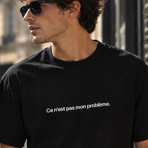 May include: A black short-sleeved t-shirt featuring the white text "Ce n'est pas mon problème." printed across the chest. The garment has a crew neckline and a standard fit, constructed from a solid black fabric with clean, simple lines.