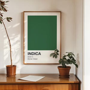 Könnte beinhalten: Ein gerahmter Kunstdruck mit einem dunkelgrünen Farbblock. Der Druck enthält das Wort "INDICA" in weißer Schrift, den Farbcode "9420 C" und den Namen "Stoner Green". Das Kunstwerk wird auf einer Holzoberfläche mit Topfpflanzen präsentiert.