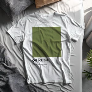 May include: White t-shirt with a large olive green square design. The text "OG KUSH" and "420 OG Ancestral Chlorophyll" are printed below the square. The shirt has a crew neck and is made of a soft fabric.