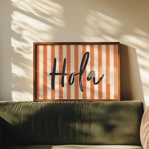 Op de afbeelding: Een ingelijste print met het woord "Hola" in zwart schrift. De achtergrond heeft verticale oranje en witte strepen. De lijst is donkerbruin. Het kunstwerk wordt boven een groene bank weergegeven.
