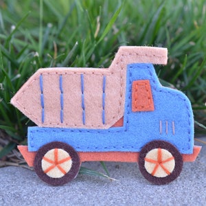 Könnte beinhalten: Ein handgefertigter Spielzeug-LKW aus Filz in Blau, Beige, Orange und Braun. Der LKW hat gestickte Details, darunter blaue Linien auf der beigen Ladefläche und orangefarbene Räder mit sternförmigen Speichen. Das Spielzeug befindet sich vor einem Hintergrund aus grünem Gras und einer grauen Oberfläche.