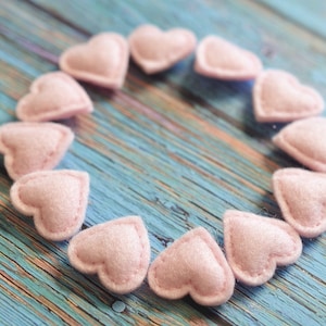 May include: A collection of light pink felt hearts arranged in a circle on a weathered, blue wooden surface. Each heart is stitched around the edges, creating a textured appearance. The hearts are evenly spaced, forming a decorative pattern.