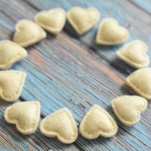 May include: A collection of small, cream-colored felt hearts arranged in a circular pattern. Each heart is stitched around the edges, creating a defined shape. The hearts are set against a weathered, blue and brown wooden surface.
